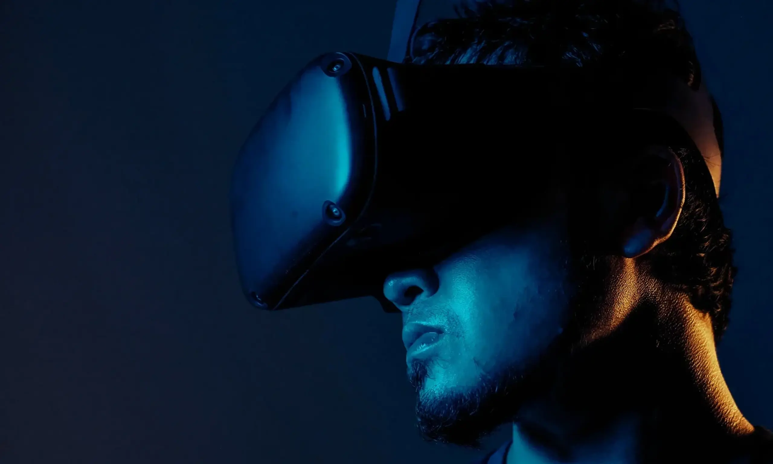 A man with short hair and a beard wearing a virtual reality headset, illuminated with blue light against a dark background.