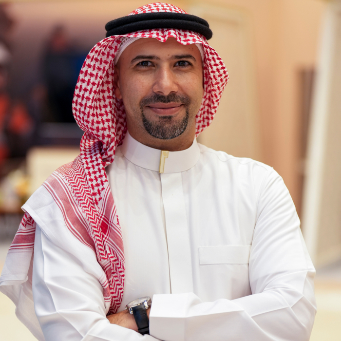 Man with a beard wearing a white thobe and a red and white keffiyeh, smiling with arms crossed.