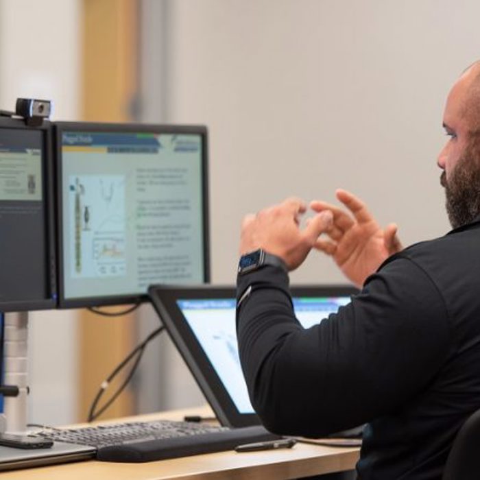 A person sitting at a desk with dual monitors and a tablet, gesturing with their hands, possibly during an online meeting or presentation.