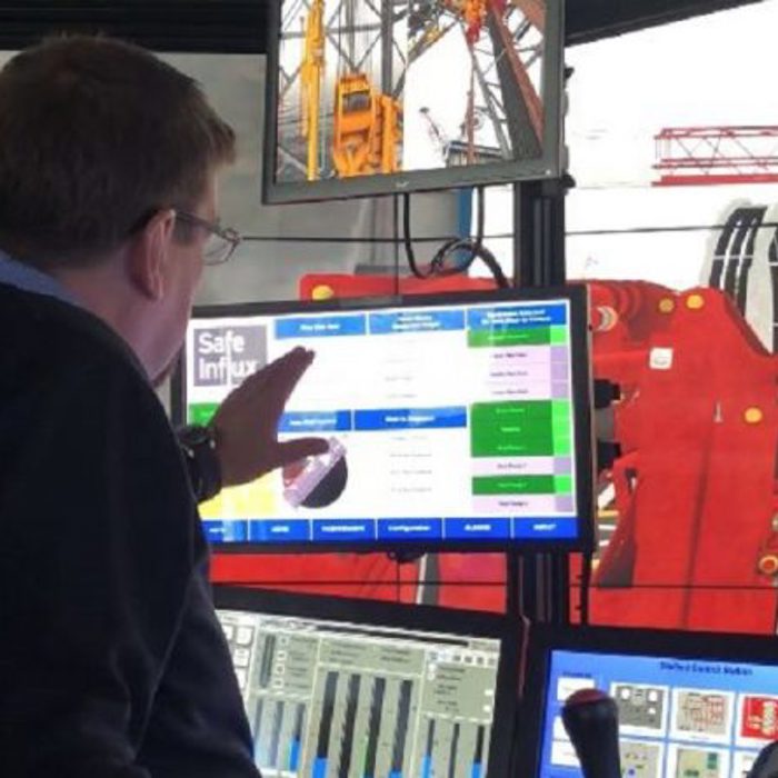 A man is sitting at a control panel with multiple monitors, gesturing towards one of them. A large piece of red industrial equipment is visible through a window.