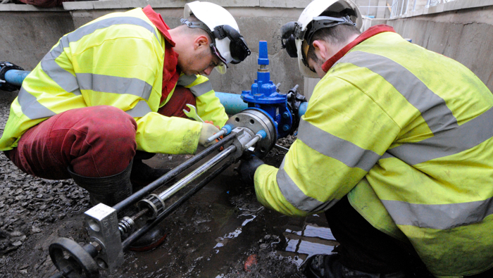 Two workers in high-visibility jackets and hard hats are meticulously inspecting and fixing a blue pipe joint in a construction area, demonstrating their training expertise.