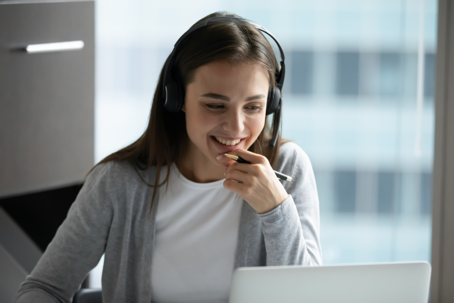 A woman wearing headphones smiles while looking at a laptop screen, engaged in an online training session. She holds a pen, appearing focused and attentive in her office setting.