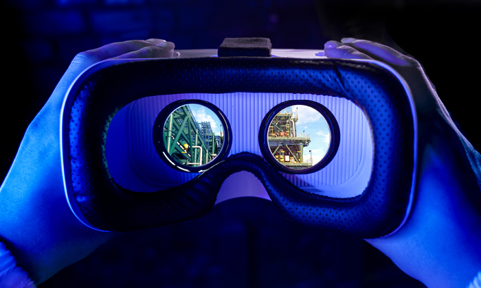 Hands holding a virtual reality headset with a view of industrial structures seen through the lenses.