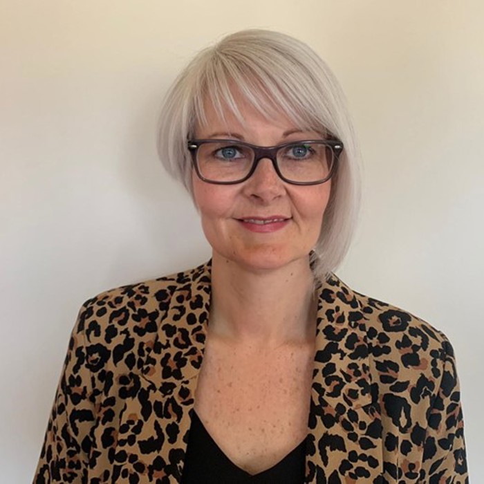 A person with short, light hair and glasses wearing a leopard-print jacket and a black top, standing against a plain background.