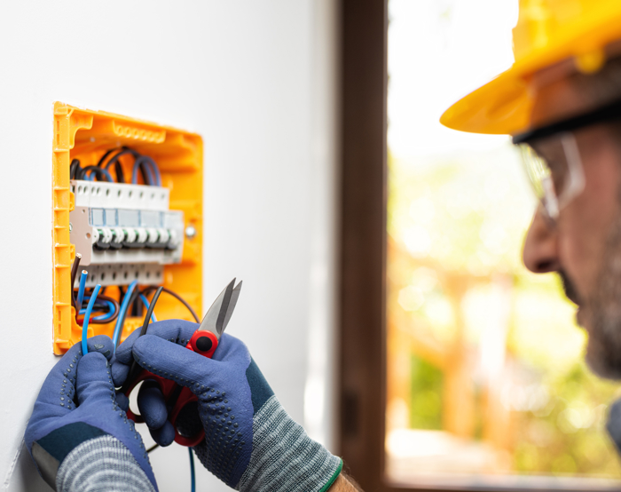 Electrician in a yellow hard hat and gloves installs a circuit breaker in a wall-mounted electrical panel.