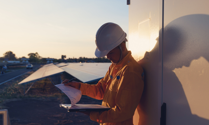 A worker in a yellow uniform and hard hat reads documents outside at sunset, leaning against a wall next to solar panels.