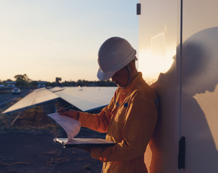 A worker in a yellow uniform and hard hat reads documents outside at sunset, leaning against a wall next to solar panels.