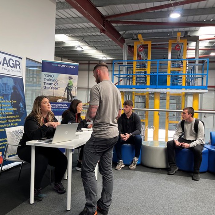 People engaging in conversation at a table in an indoor industrial setting, with company banners displayed in the background.