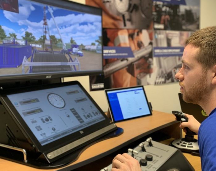 A man operates a control panel while observing a drilling simulation on two screens in a technical training room.