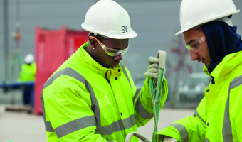 Two workers wearing high-visibility jackets and safety helmets are undergoing training as they skillfully handle a rope or strap outdoors.