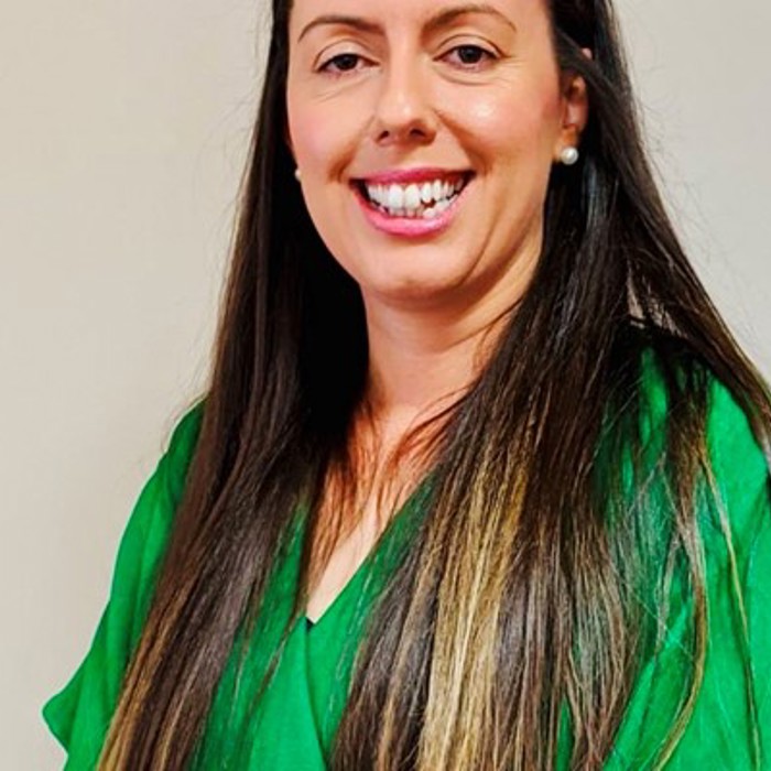 A woman with long brown hair wearing a green blouse is smiling in front of a plain background.