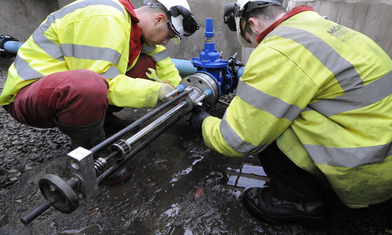 Two workers in safety gear installing a valve on a pipe in a muddy trench.